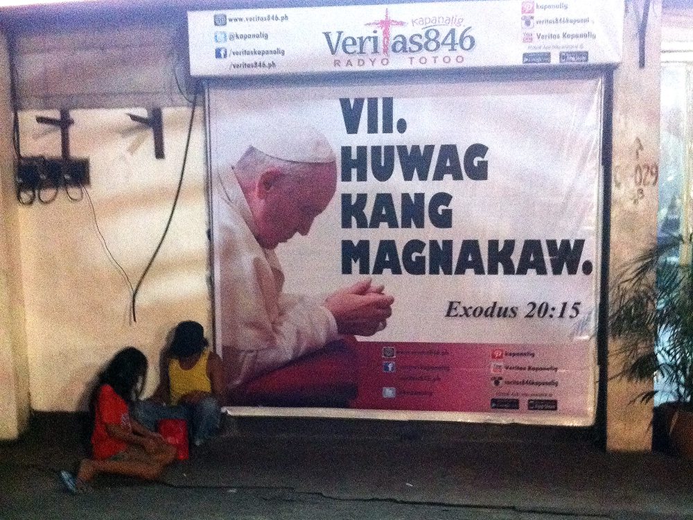 A homeless father and daughter take a break in front of an image that may just explain why there are many poor people in this country. (West Avenue corner EDSA, Quezon City)
